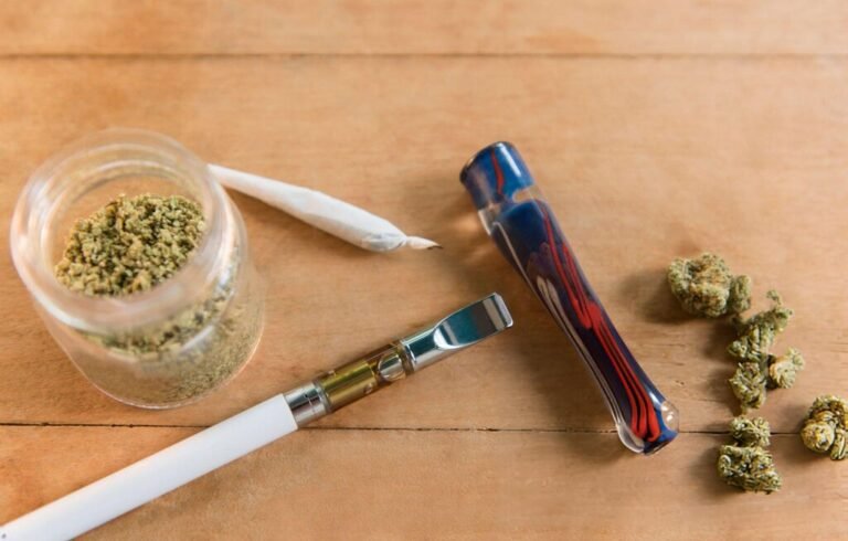 Cannabis buds, a rolling paper, a vape pen, and a glass pipe on a wooden surface.